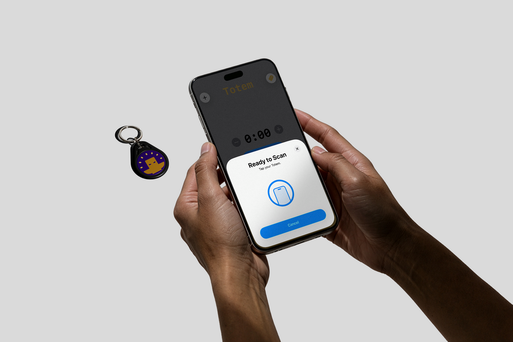 Person holding a phone with our Totem app tapping to our purple keychain Totem with a yellow logo on a gray background. Used to aid focus for ADHD and students. Totem Focus.